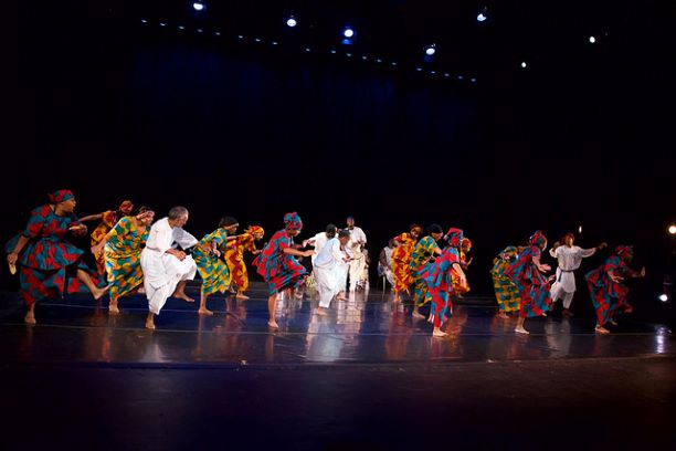 Group of performers in colorful West African garb on stage dancing West African Dance