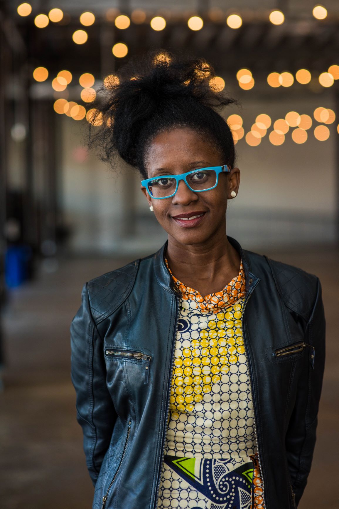 Bass poses against an illuminated background, wearing blue glasses, a multi-colored dress and leather jacket