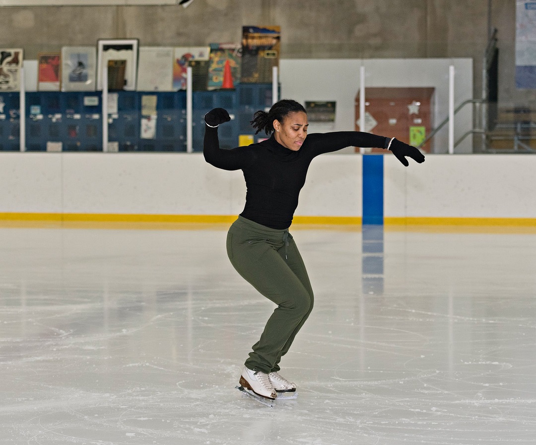 Richburg ice skates in rink wearing black top and gloves, green pants, and white ice skates