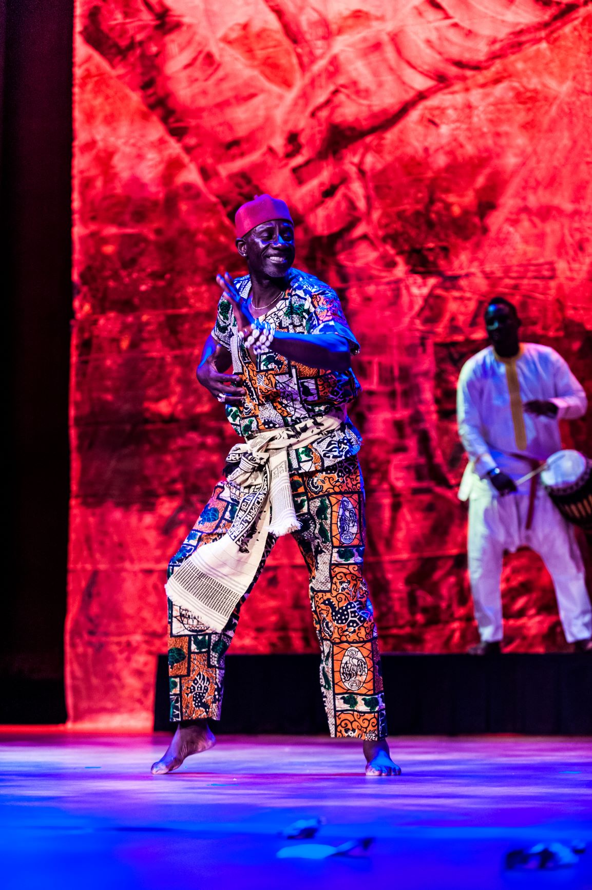 Man in multicolored African garb dancing against a red background with a drummer wearing white