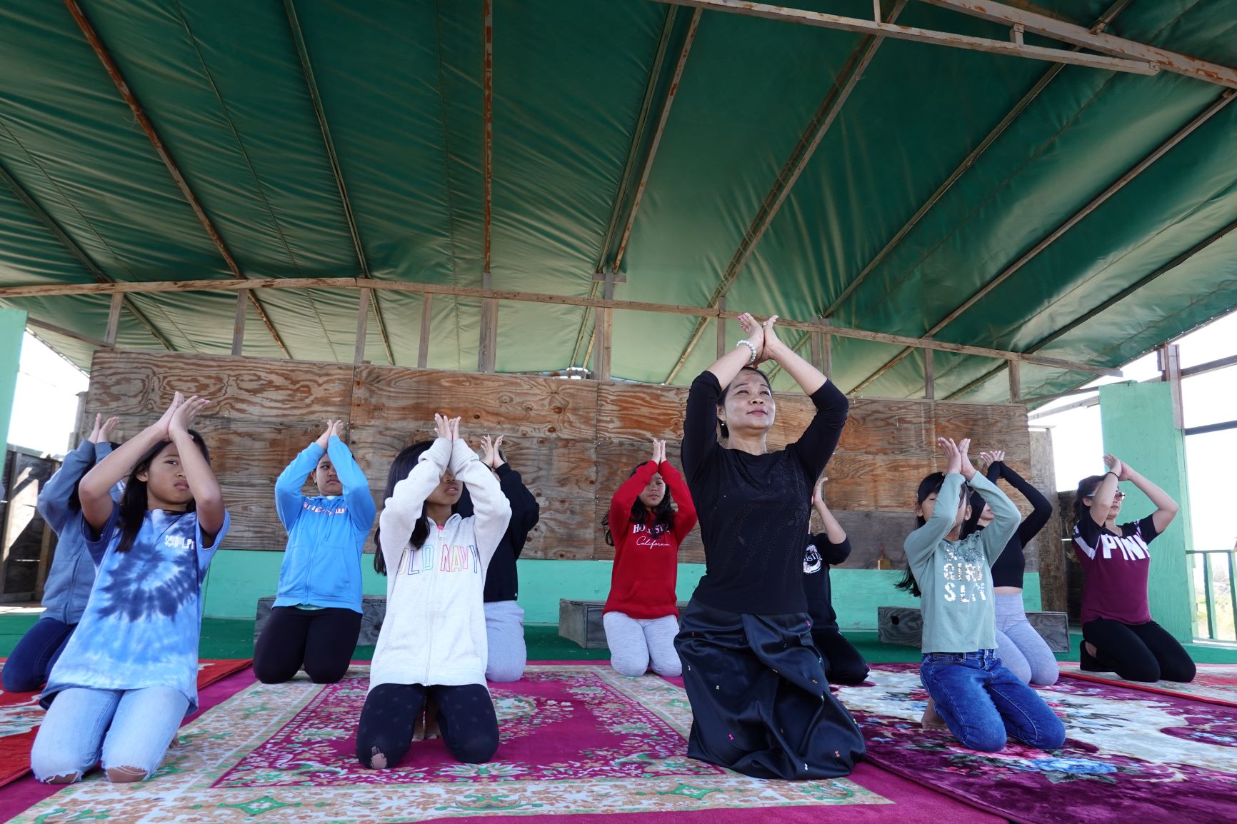Kneeling woman leading girls with arms raised, palms together at their foreheads
