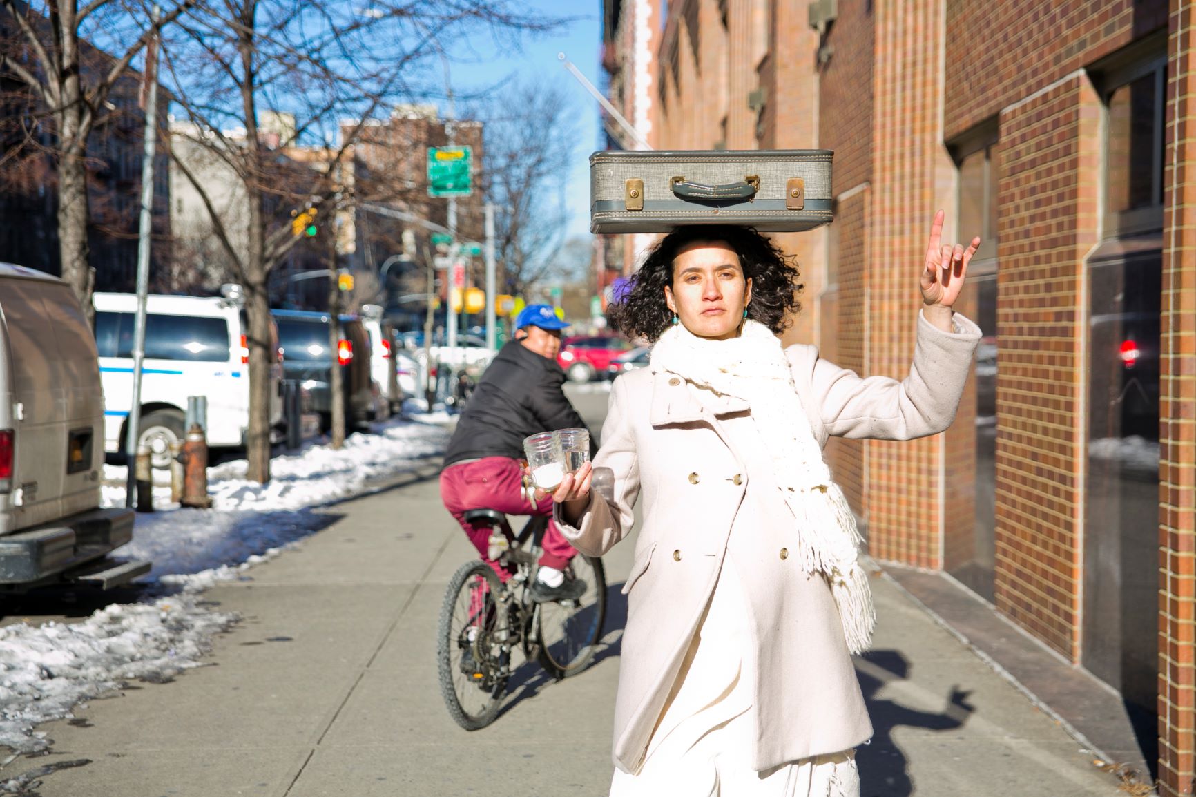 McGregor balances a suitcase on her head and carries two jars down a sidewalk with a passing bicyclist as witness