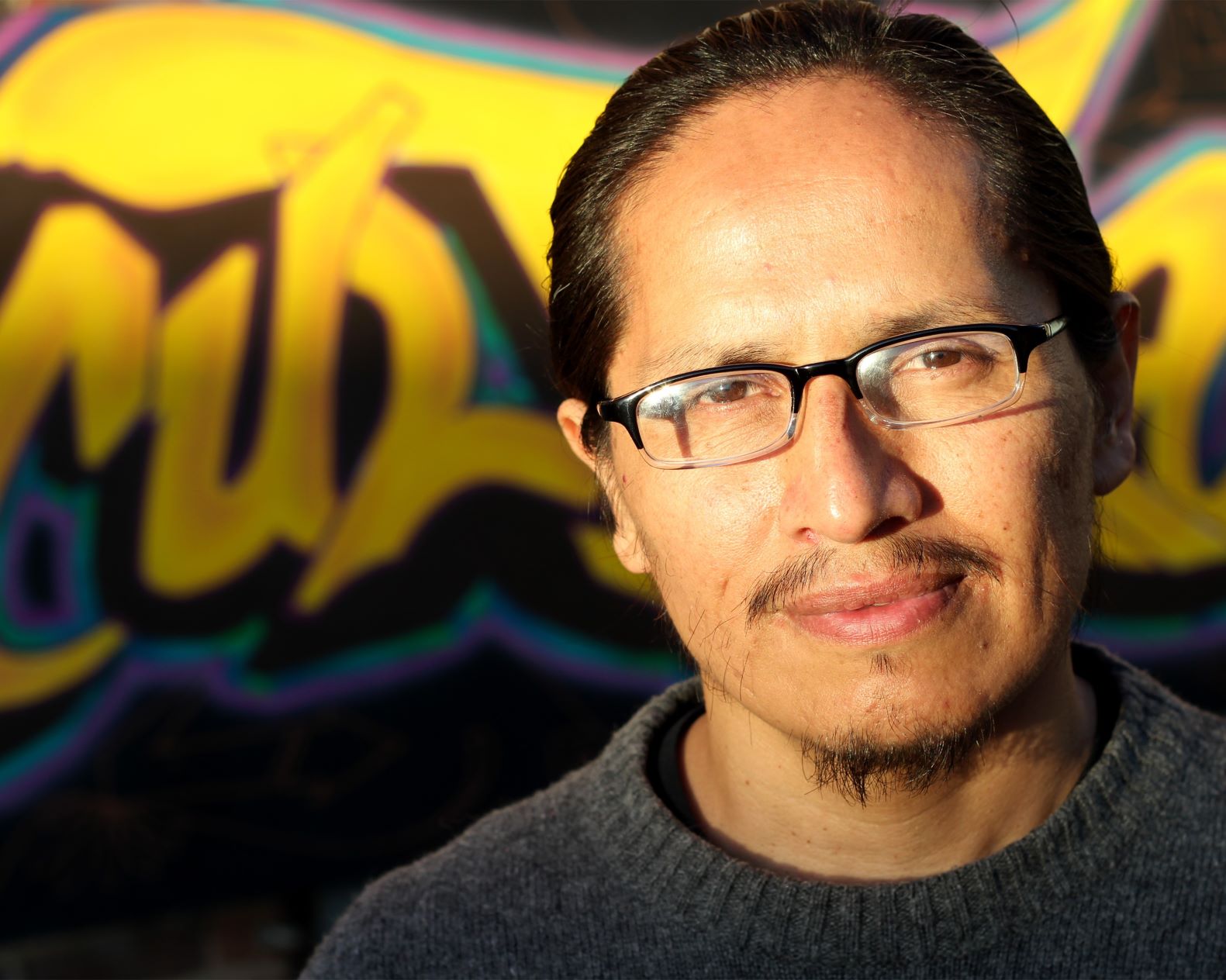Man wearing black glasses and a gray sweater poses against a street mural