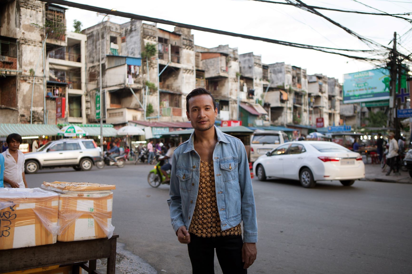 Ok poses on sidewalk of busy street, wearing a patterned shirt, denim jacket and black pants