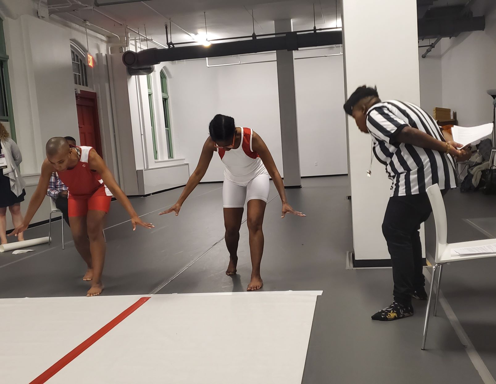 Man and woman in red-and-white biker shorts stand ready to run as referee in black-and-white stripes looks on