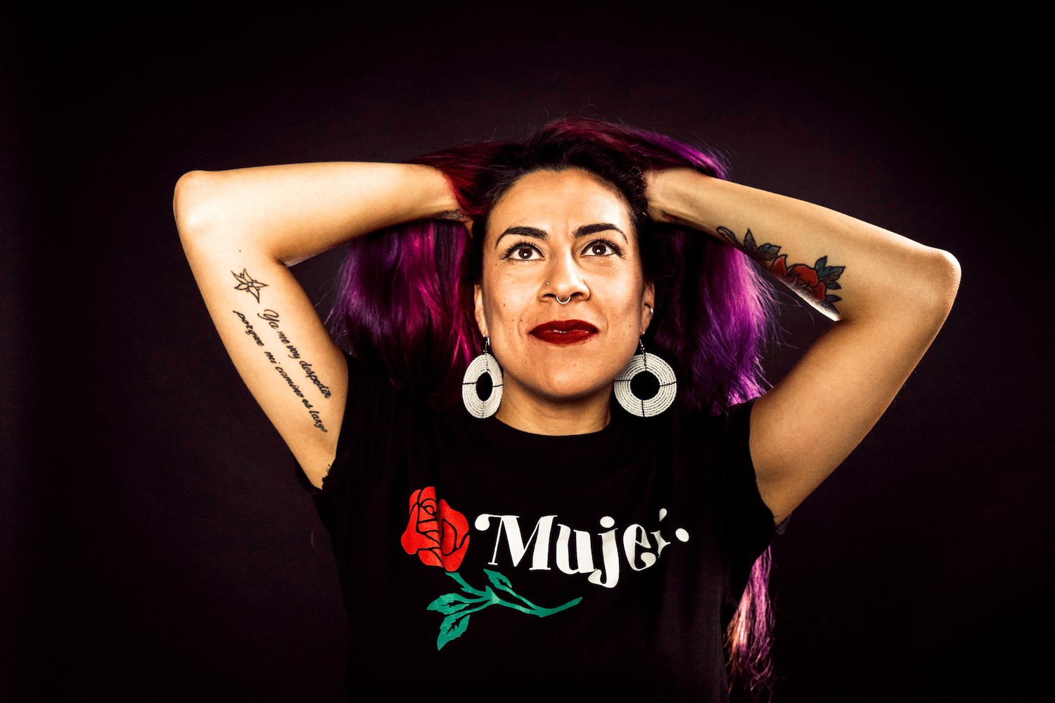 Woman against a black background with her hands in her purple hair