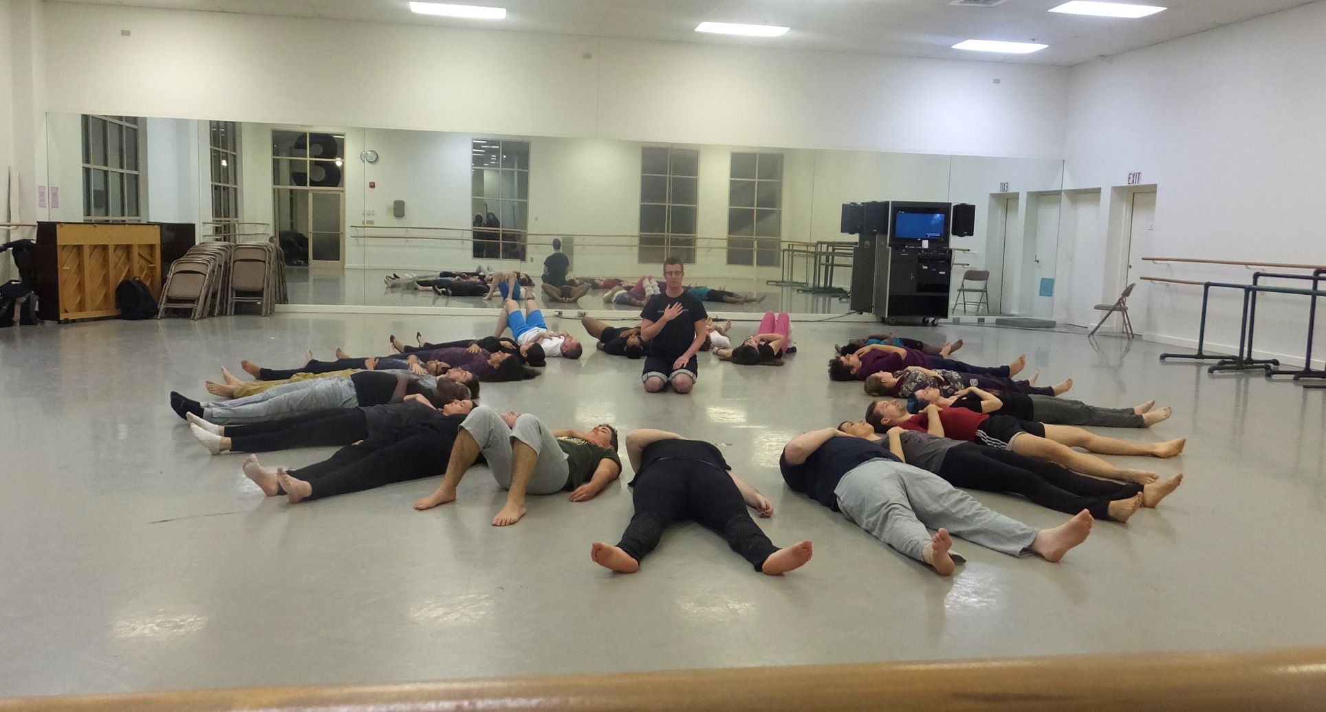 Group of people laying on their backs on the floor in a circle, heads in the middle. Dorsey sits on his knees in the center, his hand over his heart, as he speaks to the group