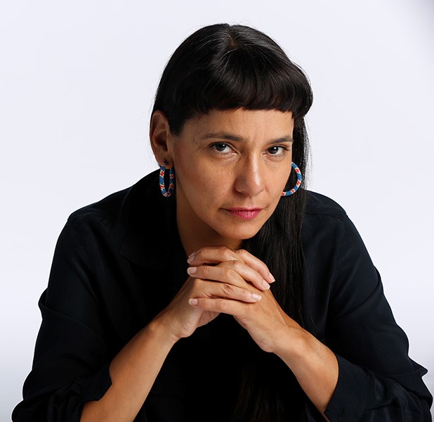 Simas, wearing a black shirt and patterned hooped earrings, poses with her hands clasped below her her chin