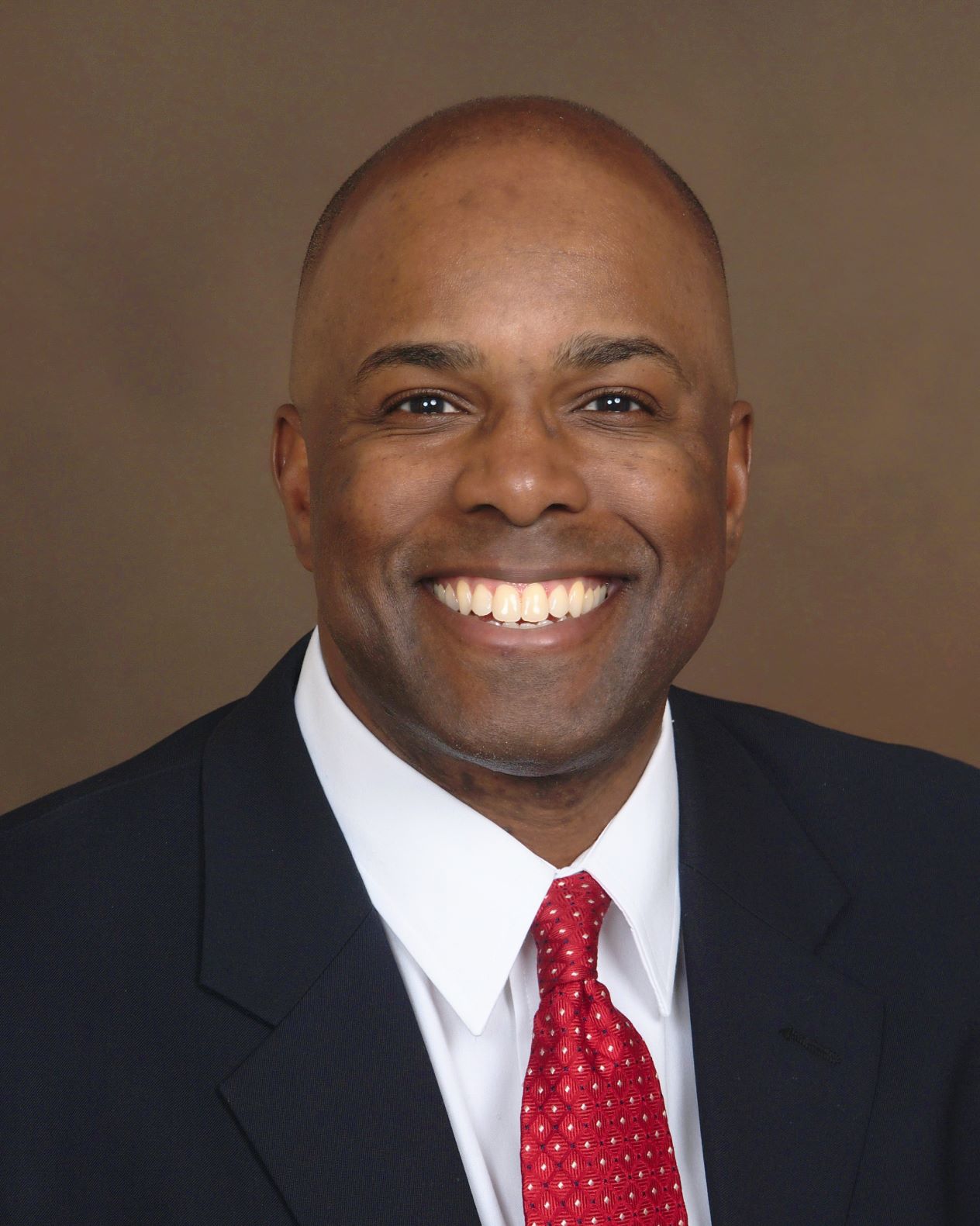 African-American man with very short hair wears blue suite, white shirt and red tie, smiles broadly