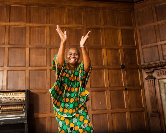 Woman in green-and-yellow outfit against a wooden background with her arms raised, about to clap her hands