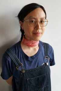 Sarah wears glasses, overalls, and a pink scarf.