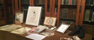 Several drawings and a pair of dance shoes on a table with a bookcase behind it