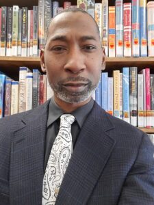 A Black man wearing a dark suit and light tie stands in front of bookshelves.