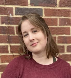 A white woman with short brown hair wearing a maroon sweater stands in front of a brick wall.