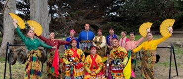 A group of 12 people in colorful garb with large fans standing outdoors on the grass in front of trees.