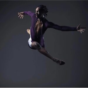 A Black man in a purple costume leaps with arms outspread, facing left.