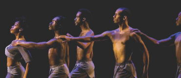 Five Black dancers stand in a line, facing left, with arms extended, against a dark background.