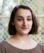 A white woman with shoulder length brown hair and a gray top in front of a blurred white and green background.