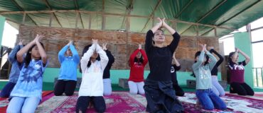 A woman and a group of children kneel on the floor performing a dance gesture