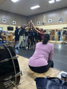 A group of dancers in a studio with their hands raised and joined and a person in a purple top with their back to the camera watching.