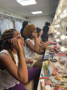 Dancers sitting in a row at a mirror doing their makeup.