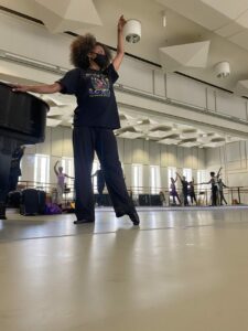 A Black woman holding a piano and lifting her arm in the air. She extends her foot in front of her in a tendu, or point.