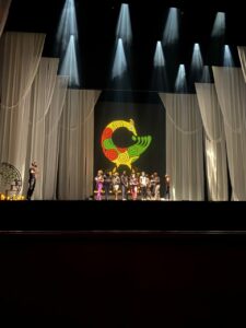 A distant scene of dancers standing in a group on stage with a figure stage right alone. A green, red, and yellow projection behind them and large white curtains frame the stage.