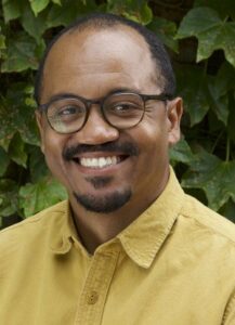 Individual with glasses, a mustache, and goatee, wearing a yellow shirt is facing the camera, smiling.