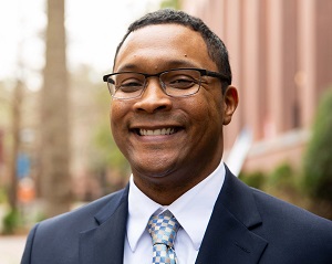 Individual with short dark hair and glasses is wearing a dark suit jacket, white shirt, and light tie, facing the camera and smiling.