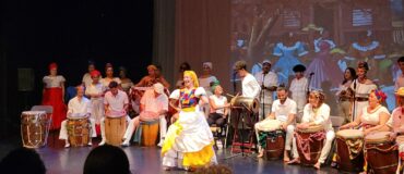 A large band of drummers and other instrumentalists behind a dancer who smiles on stage