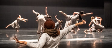 An individual stands at the front of the stage gesturing widely while on stage many dancers are moving.