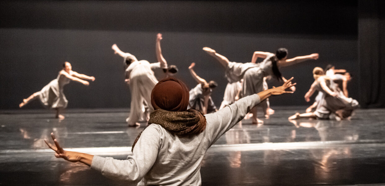 An individual stands at the front of the stage gesturing widely while on stage many dancers are moving.