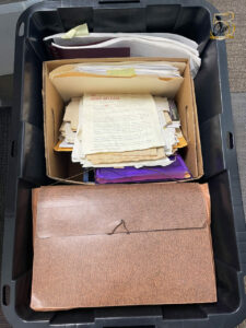 A black plastic tub filled with papers, a document box of loose materials, and a brown accordion folder