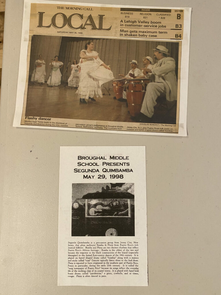 On top, a newspaper with LOCAL at the top and a large image of a dancer and drummers. Below, a white program with text reading "Broughal Middle School presents Segunda Quimbamba, May 29, 1998"