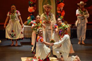 Many people on stage wearing white costumes with colorful flowers and adornments. A man and a woman kneel in front holding hands with other dancers all around them.