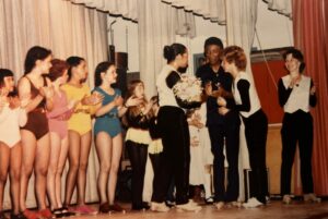 A row of children on stage in colorful leotards watching older dancers holding a bouquet