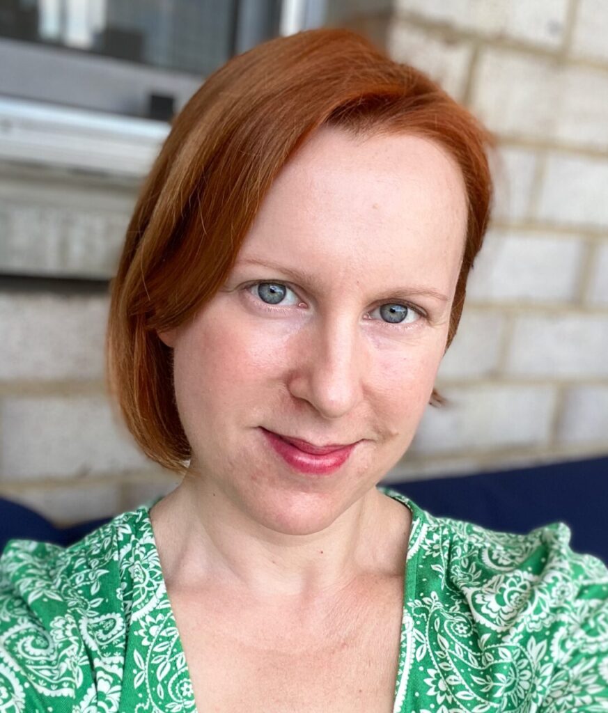 Sara Nash, a disabled white woman with chin length red hair, is seated in front of a white brick wall and window. Her head is tilted slightly to the right, and she is looking at the camera and smiling. She has blue eyes and wears red lipstick and a green and white patterned shirt.