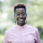 MK Abadoo, a medium brown-skinned African American person smiles. They wear a pink buttoned up shirt against a forest green background. Her black hair is in a short, tapered cut with red highlights and slender black earrings frame their wide smile. Photo by Shaka Shot Dat.