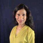Mesma Belsaré, an Indian classical dancer is seated against a dark blue background. She wears a green sequined shirt and earrings. Her hair is long, dark and untied, as she looks and smiles directly at the camera. Photo by Paul Herb.
