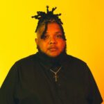 A Black man with hair styled in short twists and a beard. He wears a black short sleeve button up shirt and a gold cross on a chain around his neck. He and the background are drenched in yellow light. Photo by Spencer Hopkins.