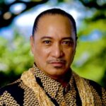 The headshot of Peter Rockford Espiritu is of a mature and handsome native Hawaiian male with black hair neatly combed back. He has brown eyes, a thin black mustache and goatee under his chin. He is wearing a traditional long sleeve, button collar, black Hawaiian print shirt in a bold geometric chevron design in a golden mustard color that resembles traditional Hawaiian Tapa or bark cloth design. Mr. Espiritu is wearing ten strands of golden brown traditional Ni’ihau shell lei that surrounds his neck and rests on the middle of his chest. He has a calm and welcoming demeanor with a slight smile on his face. Photo by Chris Rohrer.