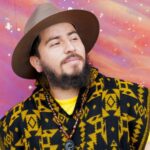 Gabriel Gutiérrez is leaning on a spray painted mural of a clouded galaxy with stars, his eyes are slightly squinted and looking toward the right of the picture. The galaxy mural is a gradient of purple, red, and orange with white dot clusters representing stars. He has dark brown hair, mustache, and somewhat curly beard and is wearing a dark brown hat, yellow shirt, black and yellowed pattern poncho hoodie. The sun is slightly shining on his slight smile, cheeks, nose and an amber necklace with obsidian dragon around his neck. His arms are going downward and slightly in frame. Photo by Leo Rivas.