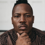Harrison Guy, a Black man is wearing a long-sleeved brown shirt with black lines at the top around the neck, across the shoulder, and down the outer chest area. His hair is cut on the sides with some left on top. His right fist touches his chin. Photo by Taylor M Hayden.