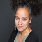Julie B. Johnson, a Black woman with light skin, and curly dark-brown hair pulled into an afro puff, smiles as she leans toward the camera with her arms crossed in front of her. She wears dangly earrings, a green blouse, and a black sweater. Photo by Patricia Villafañe.