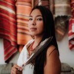 ShanDien Sonwai LaRance stands in Traditional white Native American dress. She holds a feather in her right, hand over her chest. ShanDien is a young Native American woman. She wears her hair down, straight and black. Behind her on the wall are traditional woven rugs with many colors; dominantly in red, orange, and brown colors. ShanDien is wearing a red coral necklace and turquoise inlaid bracelet. Photo by Wes Cunningham from One Trip Media.