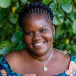 Muisi-kongo Malonga, an African woman with deep brown skin and dimples. She is pictured in front of a background of green leaves and is smiling. Her hair is braided and styled into a crown. She is wearing a silver chain with a cowrie shell pendant and gold teardrop earrings with an orange stone. Her top is a multi-colored African-print blouse with midnight blue, sea green and light peach-colored motifs. Photo: Bethanie Hines.