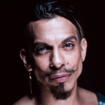 Luis Ordaz Gutiérrez wears an indigo shawl with embroidered indigenous motifs and a deep plunging V neckline that extends from one collar bone to the other. He is dramatically lit and appears to be floating out of a pitch-black background. He is a brown-skin Mexican man with a goatee beard and a styled mustache that curls up at each end. He gazes directly towards you with a slight angle of his head. Photo by TheArtist JohnnyV.