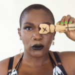 A bust length photograph of Nkeiruka against a solid white background. Her hair is close cropped and her hand is holding up an opi with an x on it covering one eye. She has on black lipstick, and gold and silver nose rings and earrings. She is wearing a black bra, and black and white graphic patterned sleeveless bra string top. Photo by Sasha Kelly