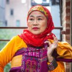 Image description: Potri Ranka Manis is wearing a colorful malong (Filipino dress) with a red scarf covering her hair. She is looking out with her hand gestured on a fire escape. Photo by Hamza Razuman.