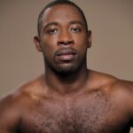 Iquail, a Black man with a buzz cut and goatee, looks directly into the camera. He is bare-chested, set against a light brown background. Photo by Rachel Neville.